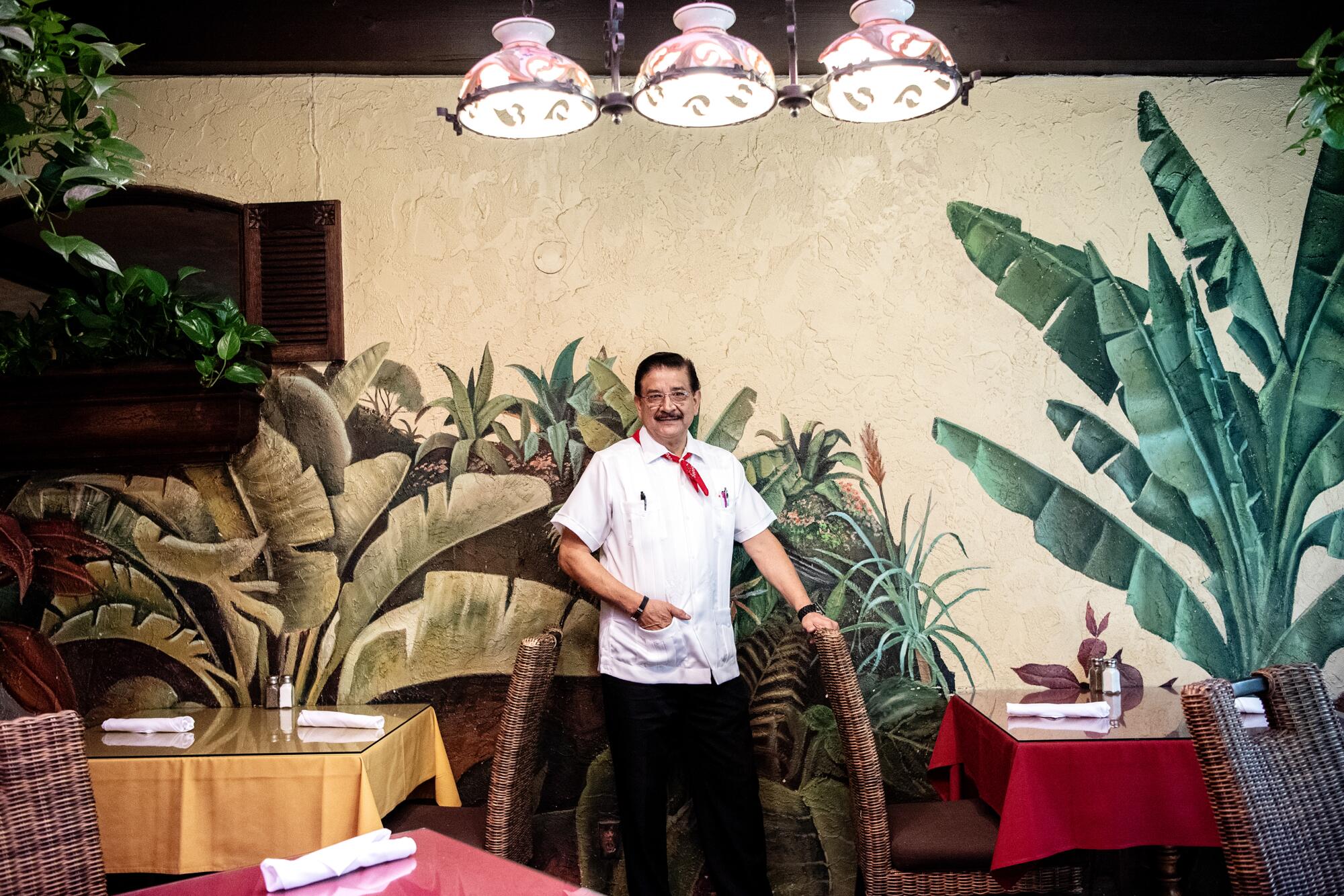 Justino Romero standing inside between two tables and in front of a wall with a colorful plant mural.