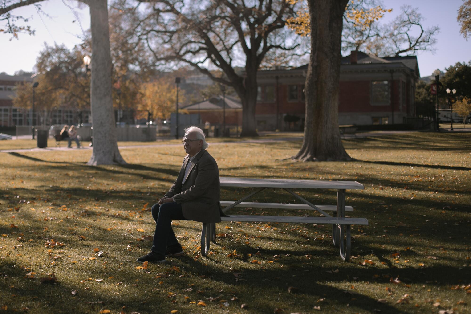 A white-haired man at a picnic table, looking into the distance as trees cast shadows across short grass littered with leaves