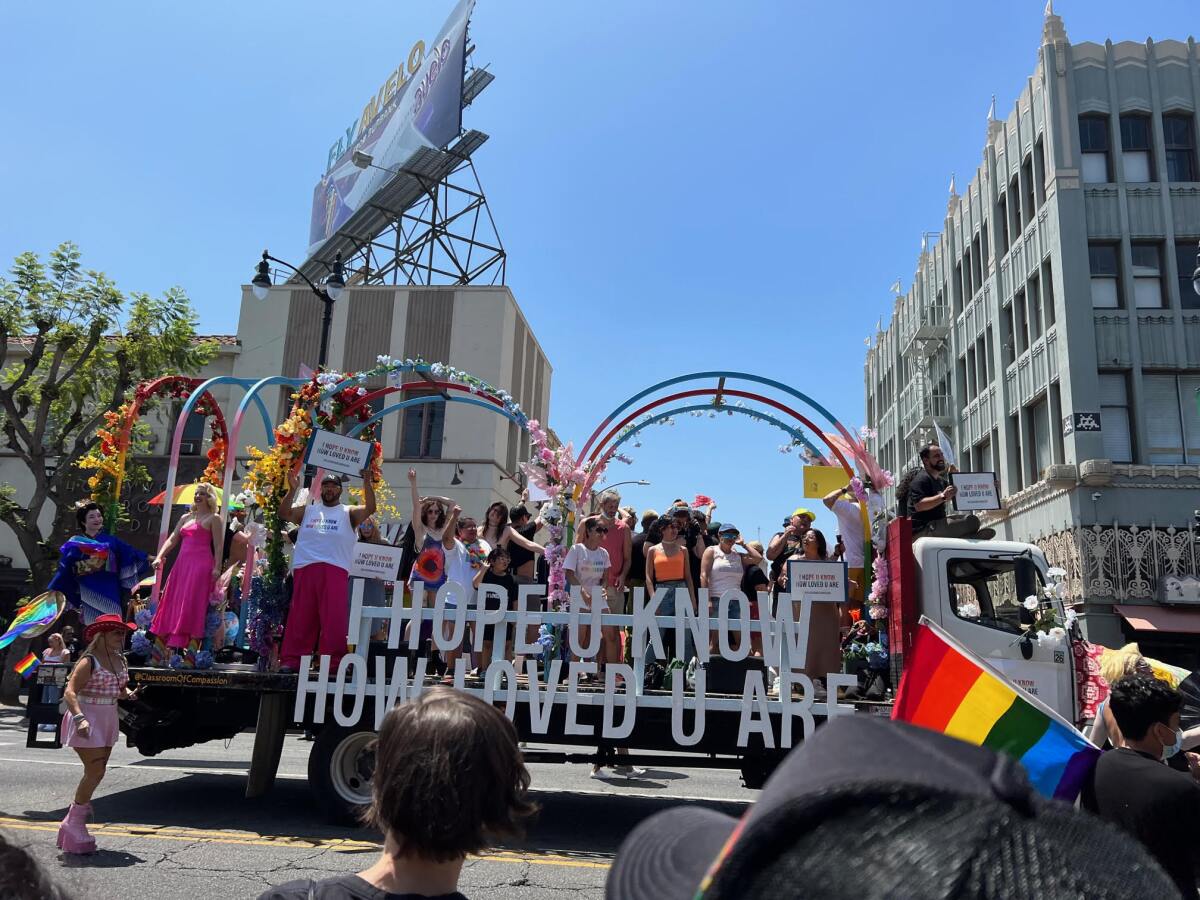 People stand atop a float, holding signs, on a city street. Amid the crowd watching is a rainbow flag.