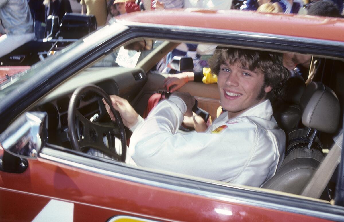  Actor John Schneider sits inside a car, looking out the window and smiling.