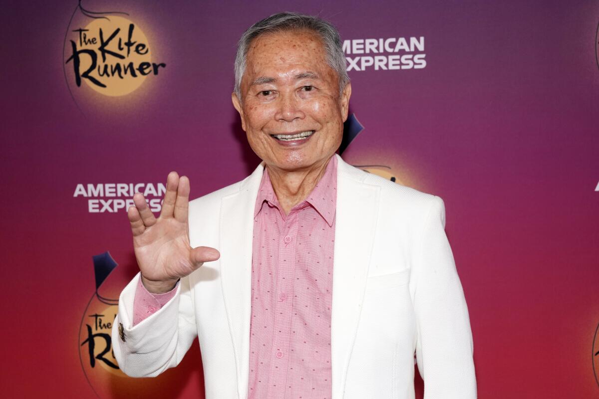 George Takei smiling, making a "live long and prosper" symbol with his hands and wearing a white suit