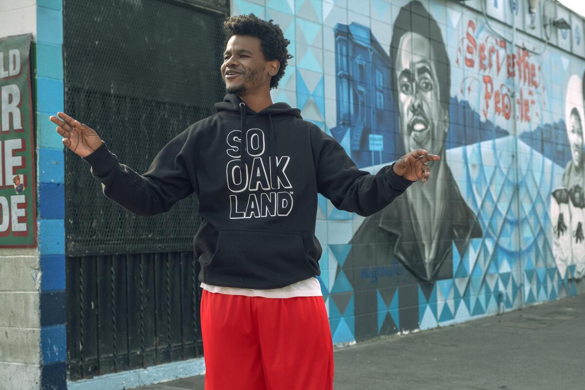 A man in a "So Oakland" hoodie standing before a mural
