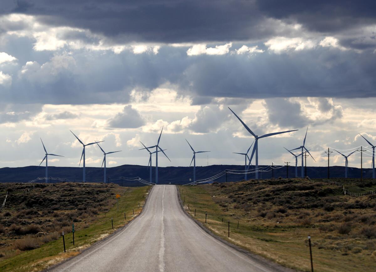 Beneath a partly cloudy sky, giant wind turbines bracket a blacktop highway in a rural area of hills and low mountains.