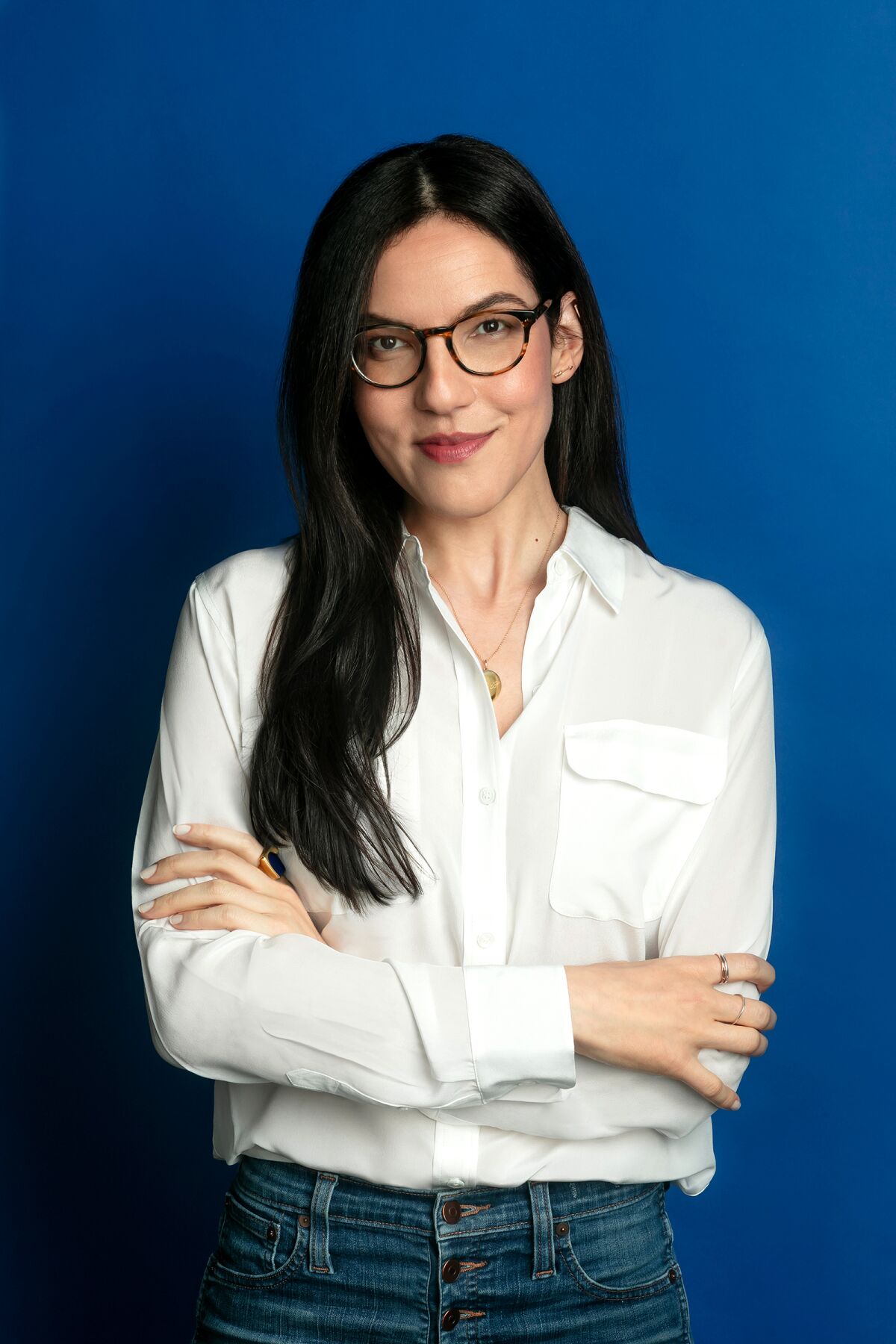 A woman in glasses, a white button-down shirt and jeans folds her arms.