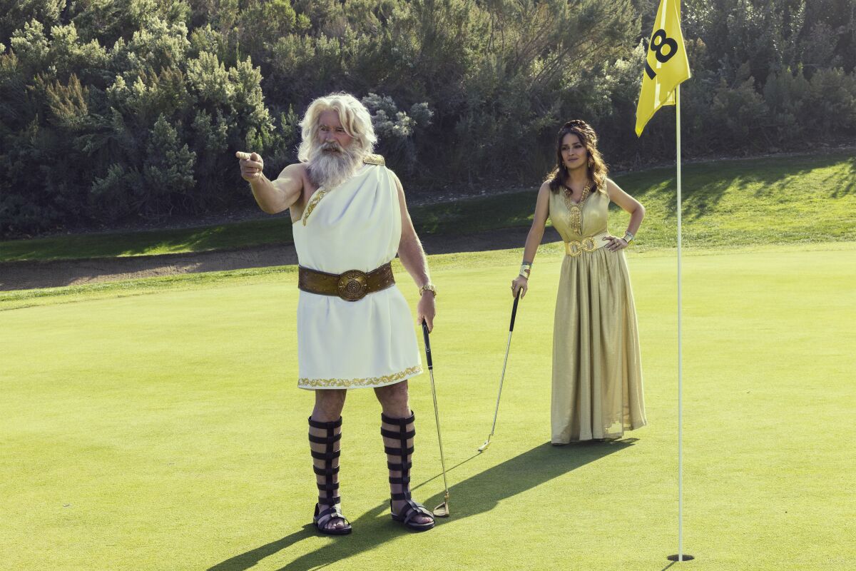 A man in gladiator sandals and a toga and a woman in a long gold dress play golf.