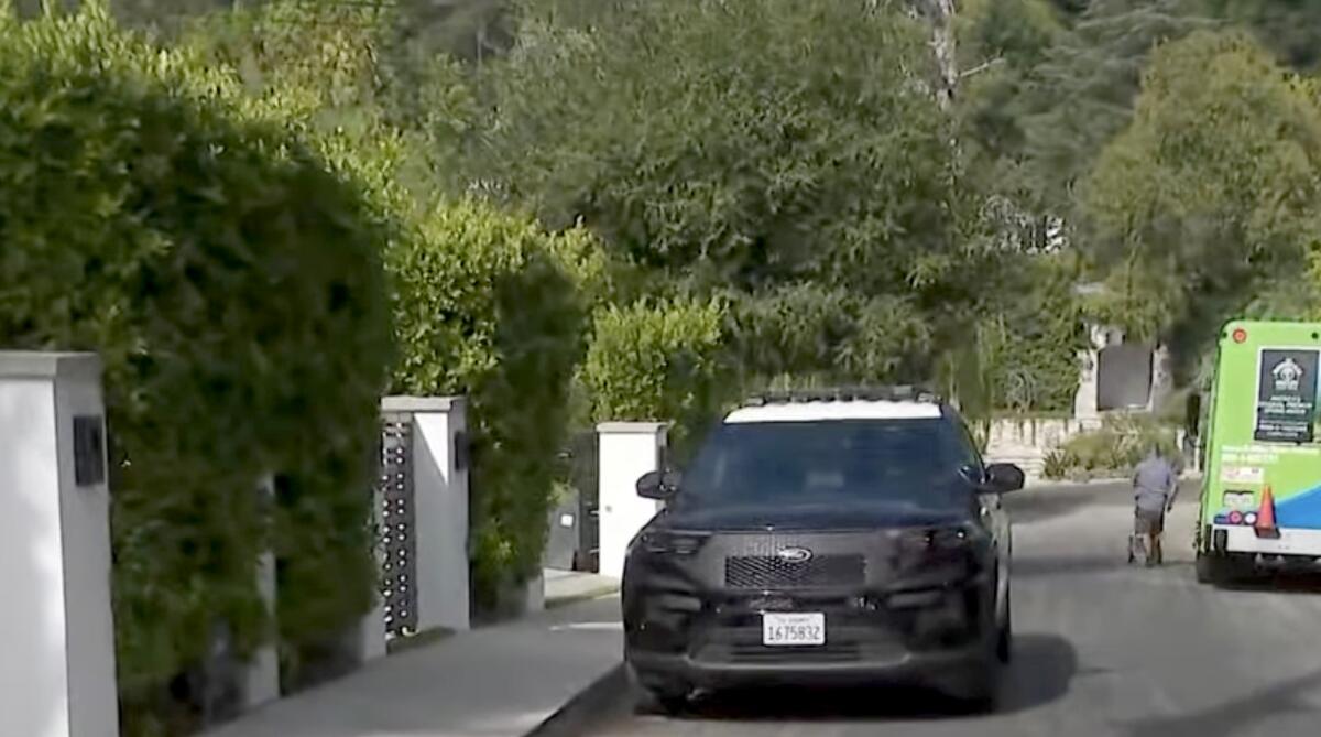 A black vehicle with a white roof alongside the sturdy white columns of a fence on a street lush with green trees and shrubs