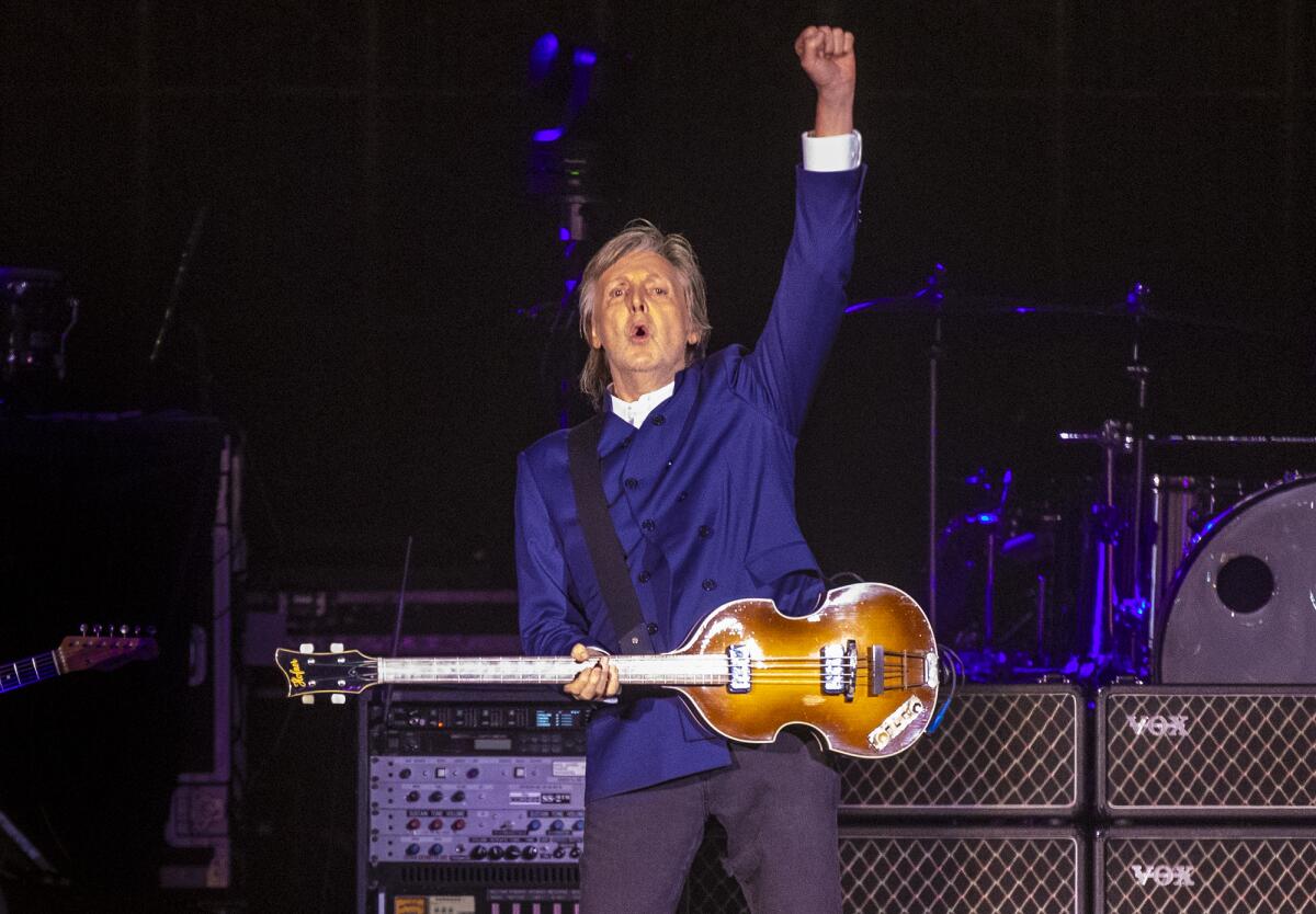 Paul McCartney in a blue long-sleeve shirt on a stage holding up his left hand in a fist, while holding a guitar