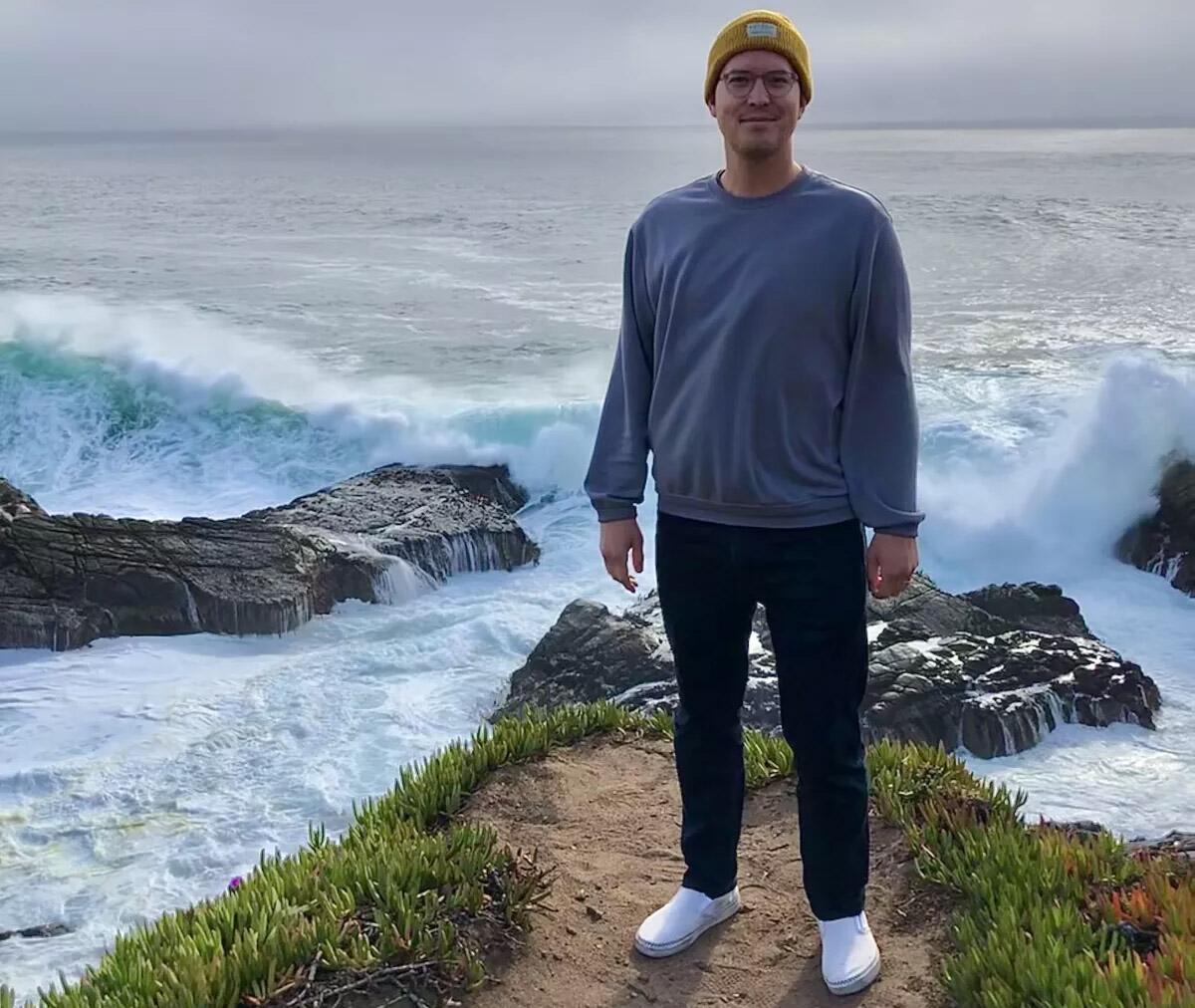 A man in a beanie and glasses stands next to the ocean where waves are crashing onto rocks.