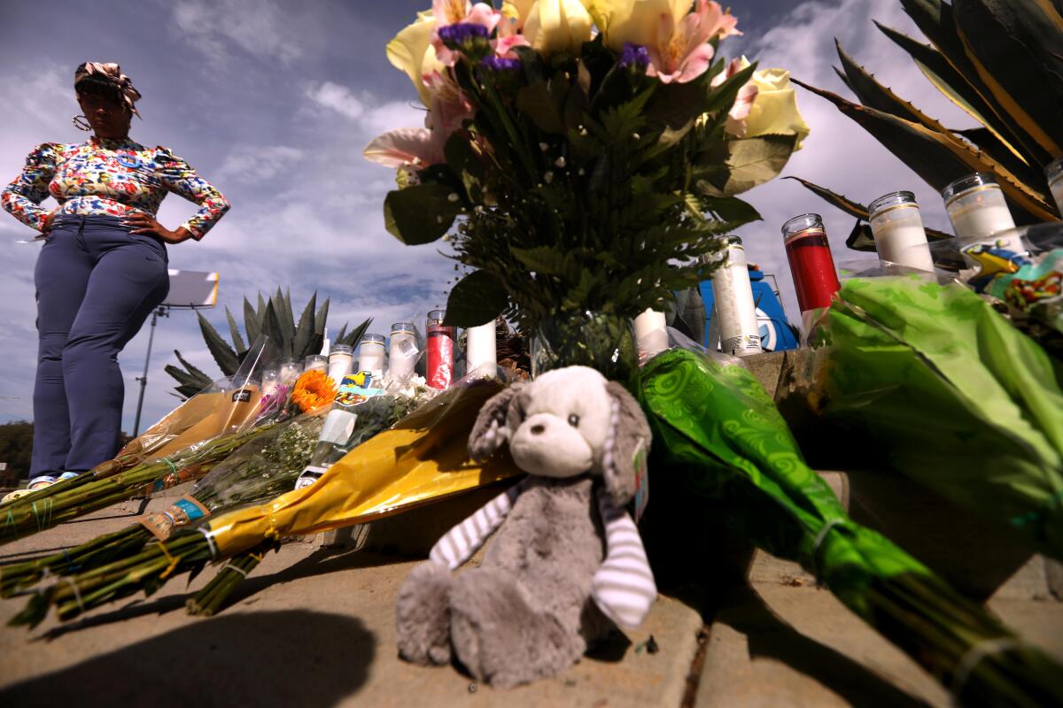 A woman looks down at flowers and candles and a stuffed animal on a sidewalk.