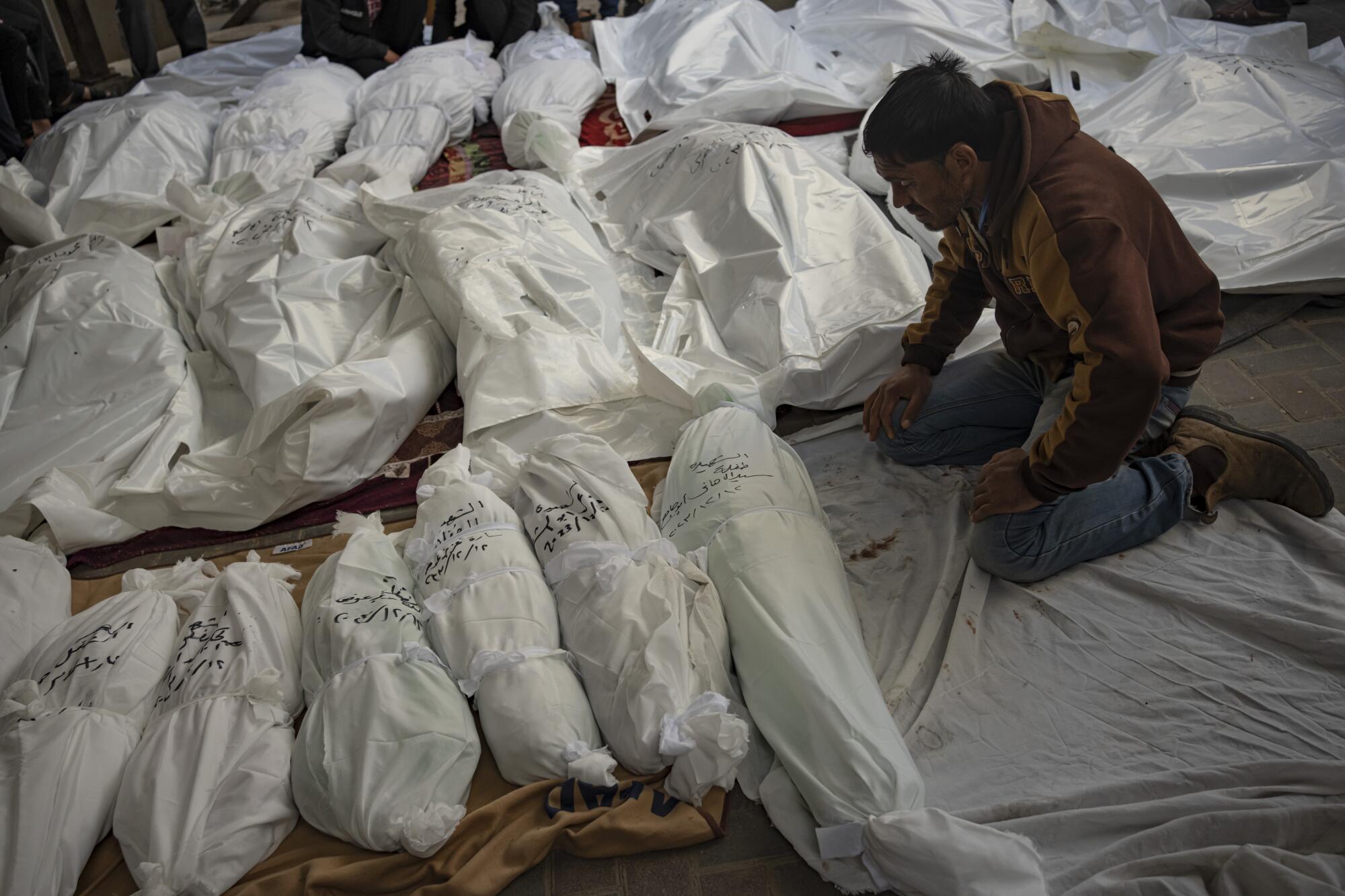 A man kneels by rows of shrouded bodies in Gaza.
