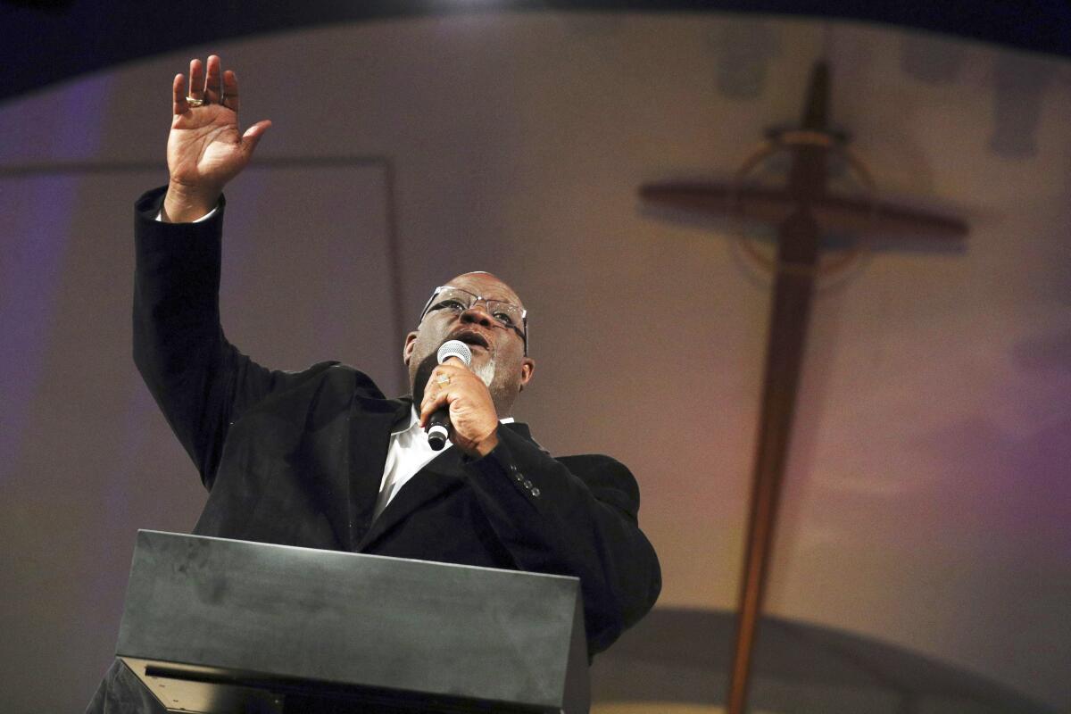 Dwight McKissic Sr. raising his hand as he speaks from a lectern, with a large wooden cross on the wall behind him.