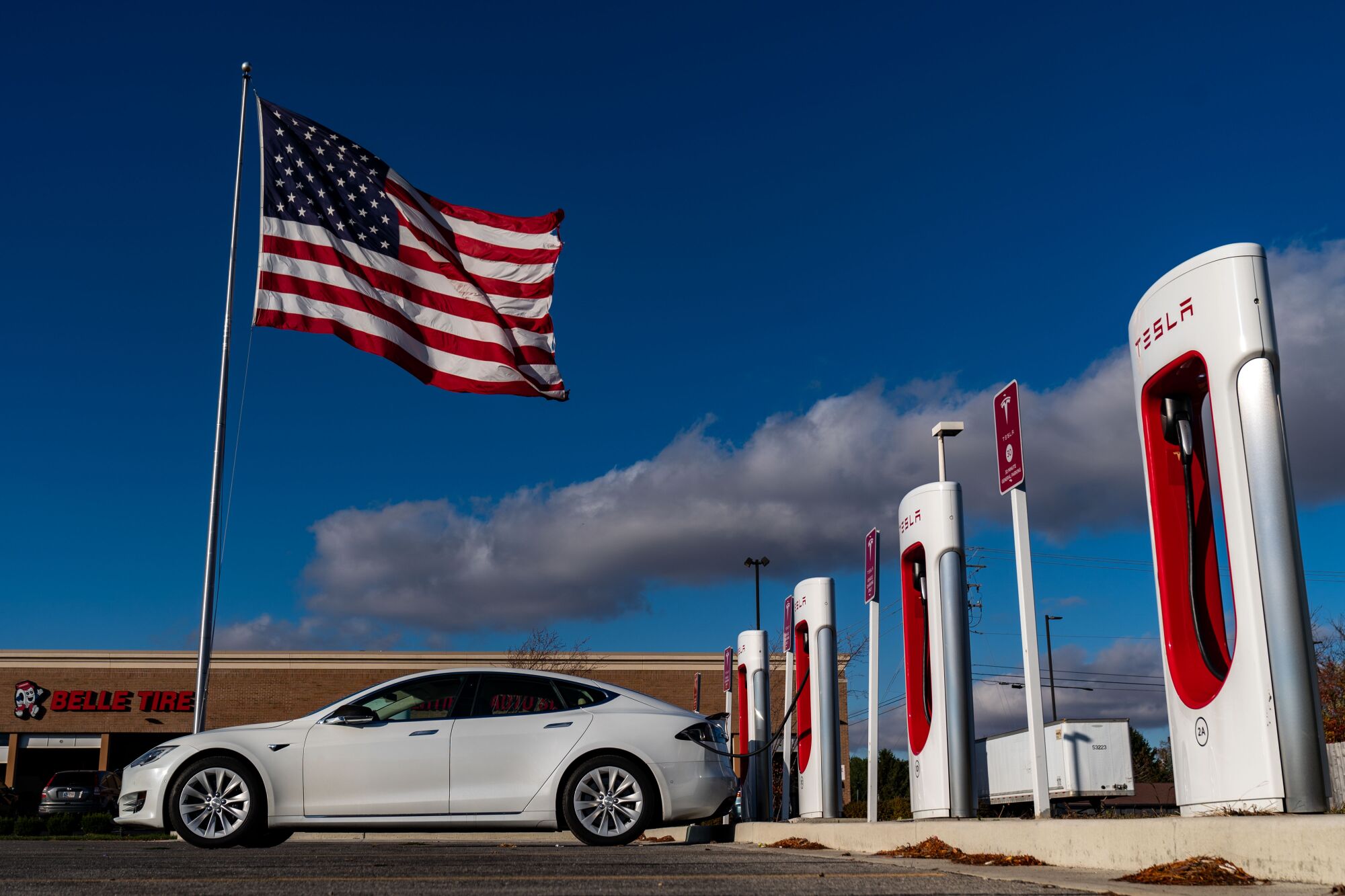 A U.S. flag flies near a white car parked in front of a row of vertical white-and-red structures with the word Tesla on them