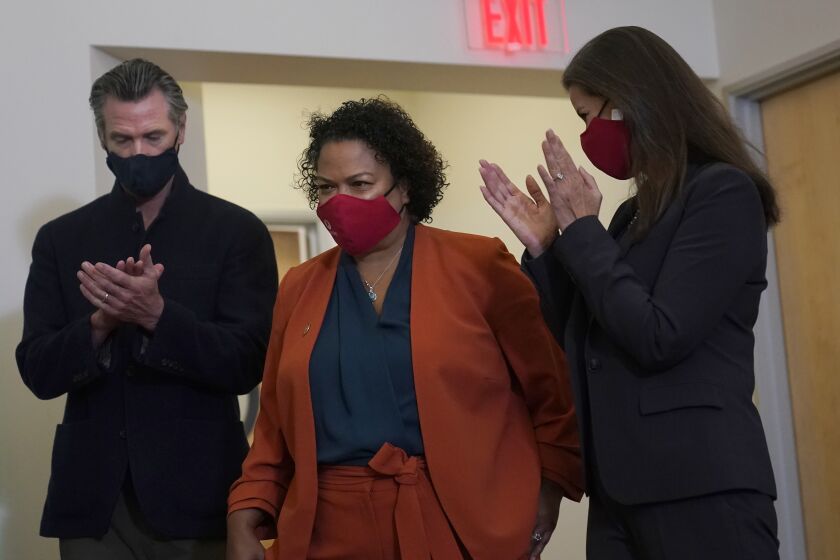 Assemblymember Mia Bonta, middle, walks to a podium to speak between Gov. Gavin Newsom, left, and Oakland Mayor Libby Schaaf