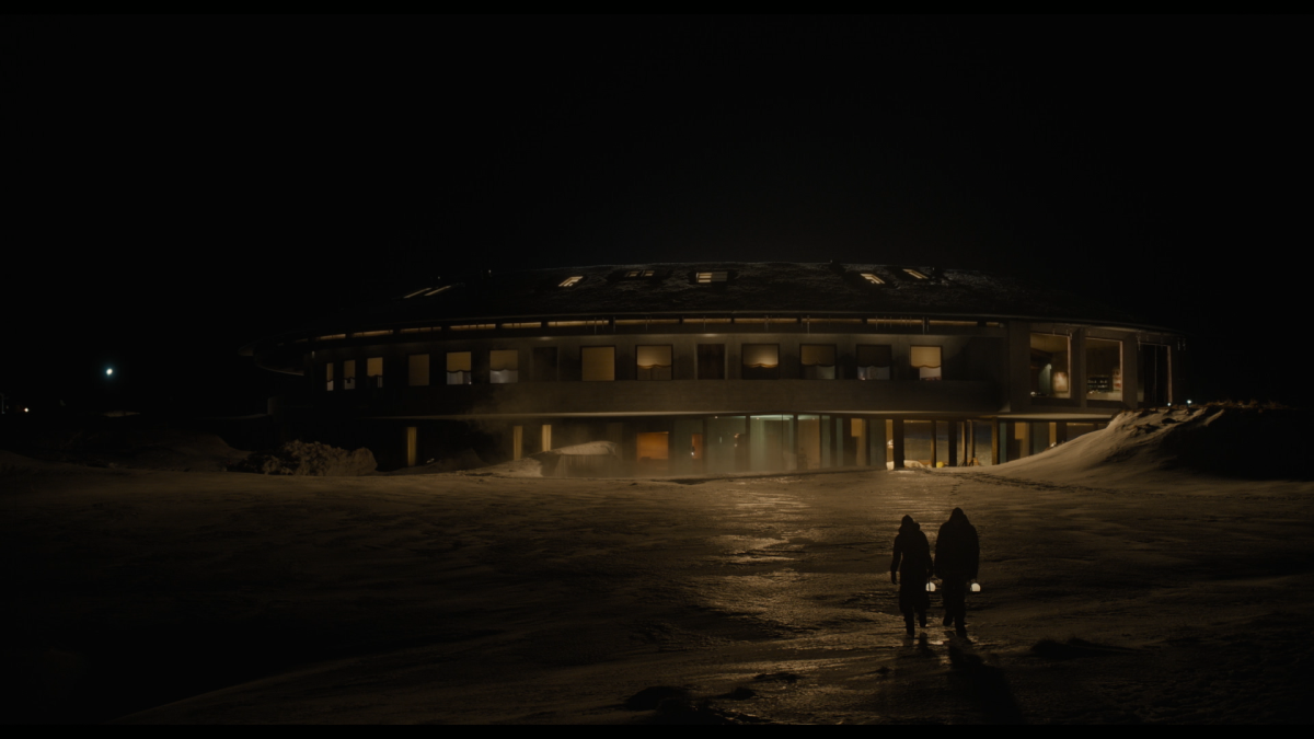 Two people walk toward a circular hotel in the snow and ice at night.