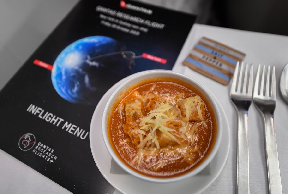A bowl of spicy tomato soup surrounded by an in-flight menu, salt and pepper packet and silverware.