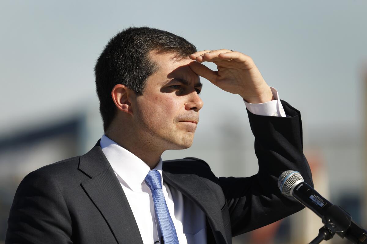 Pete Buttigieg stands at a microphone while shielding his eyes from the sun