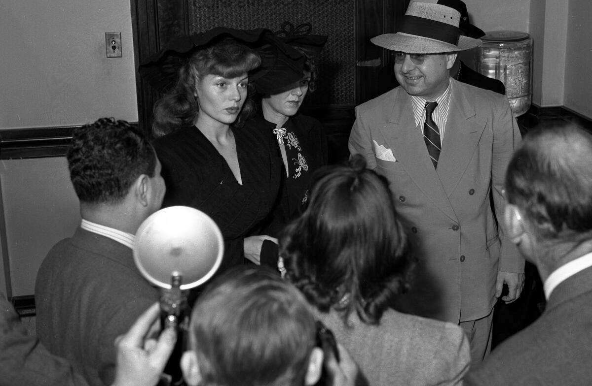 Two women are surrounded by people, including a man in a hat, one person in the crowd holds up a flash for a camera.