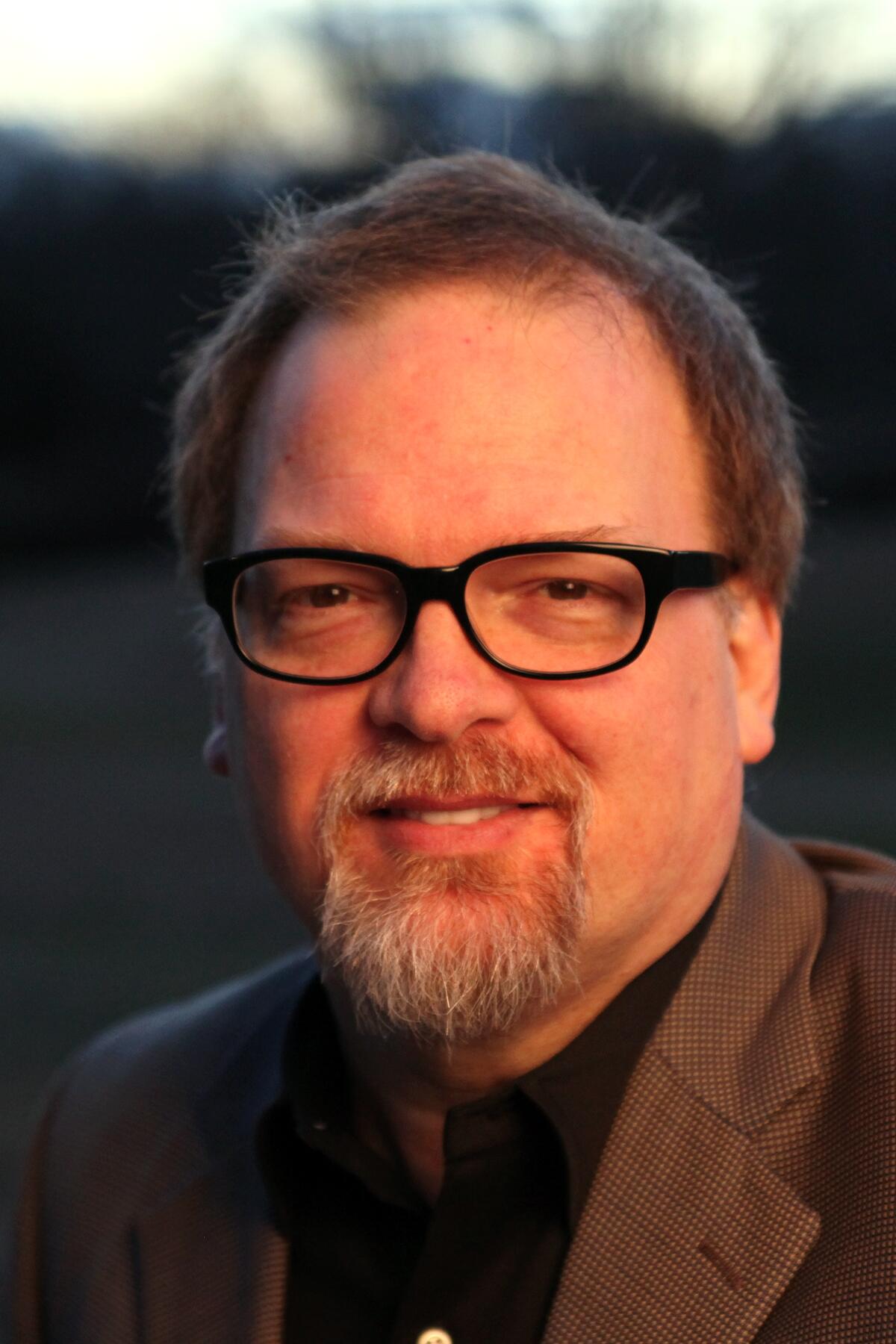 Greg Iles, in black-rimmed glasses and a goatee, smiles to the camera.
