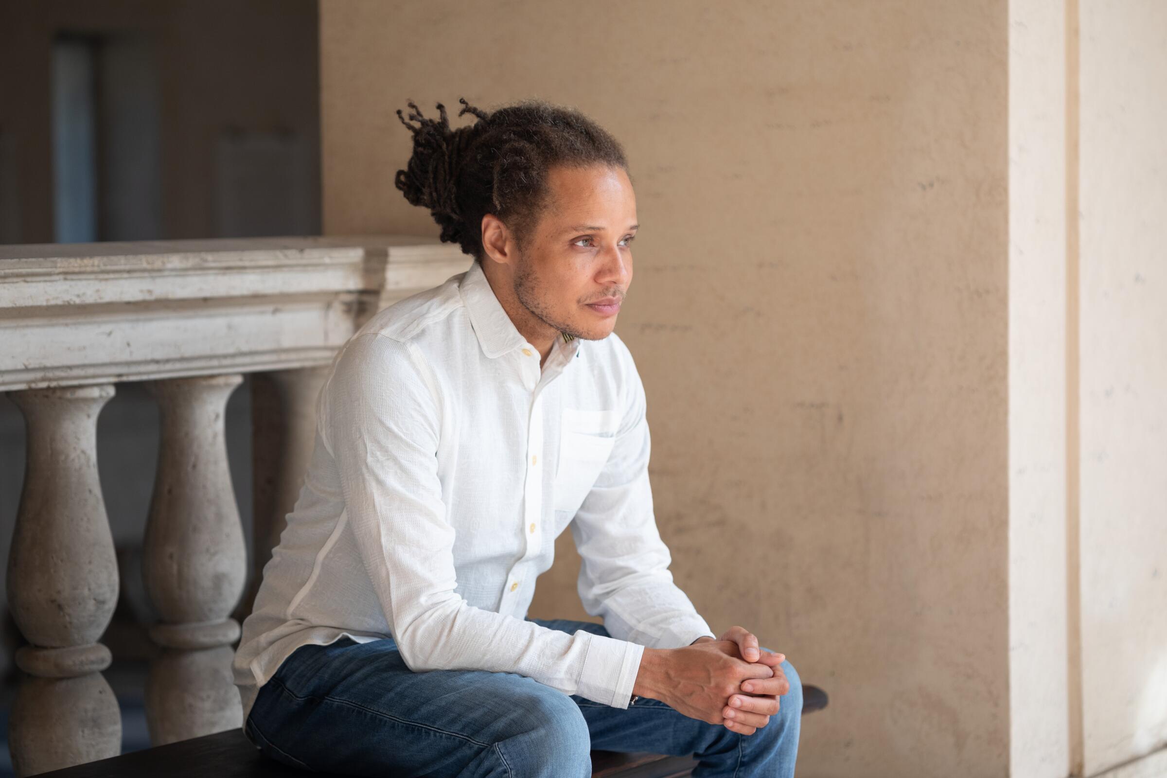 A man in a white shirt and blue pants sits contemplating.