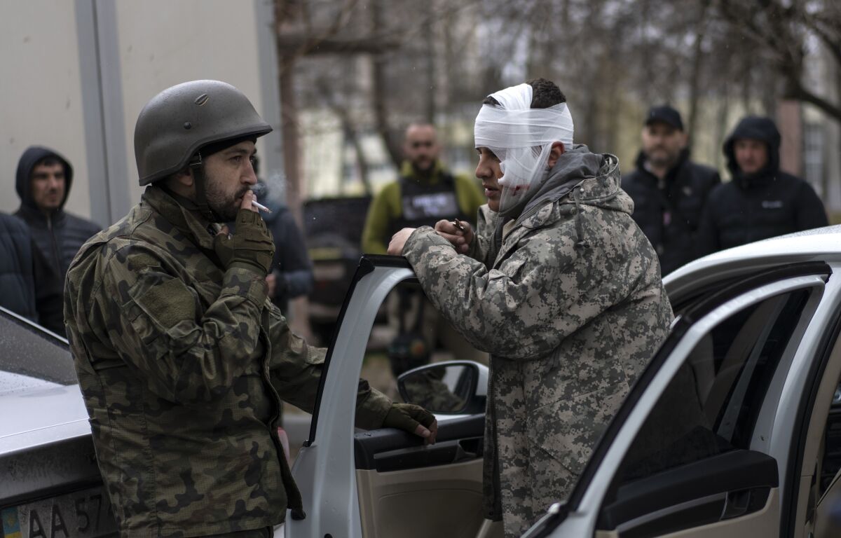 A man in fatigues, his head bandaged, right, stands by his car door as he speaks to another man in fatigues, who is smoking 