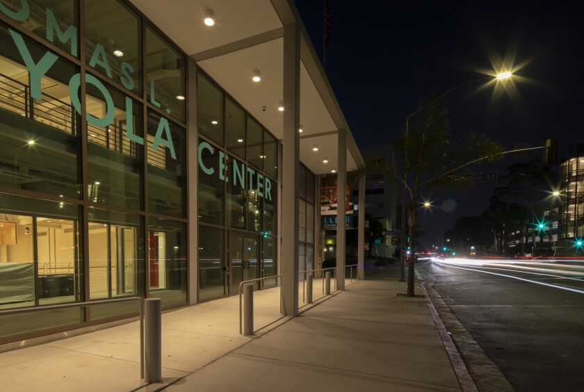 An angled view of the facade of the Beckmen YOLA Center at night illuminated from within. Traffic streams to the right.