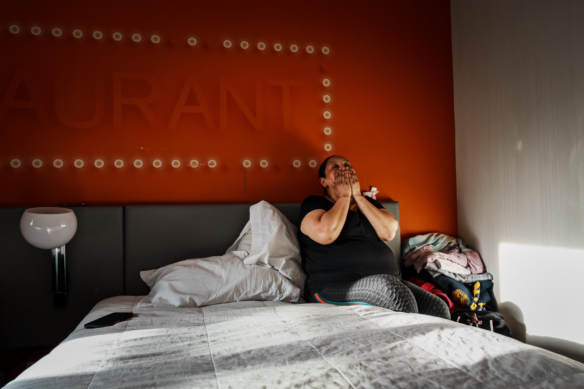 A woman puts her hands to her face while sitting on a bed. 