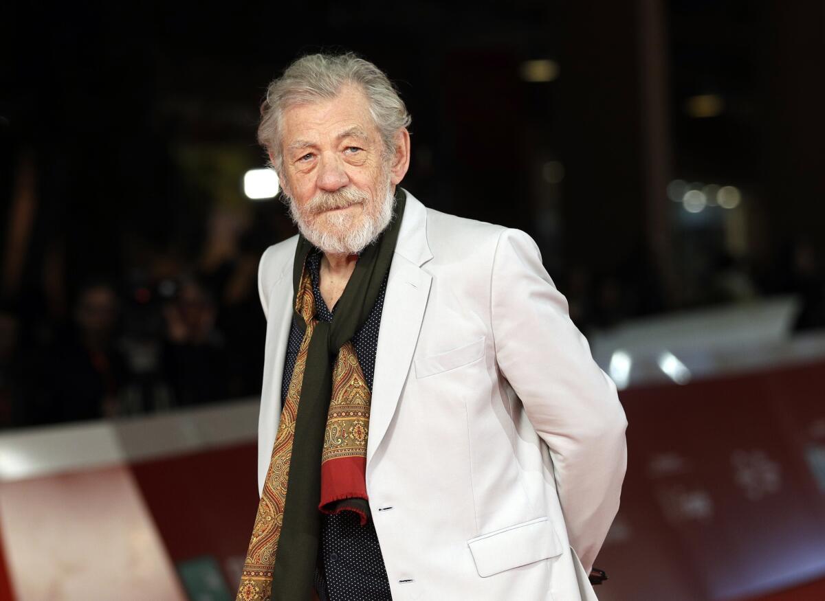 Ian McKellen, with a gray beard, dark shirt, white jacket and colorful scarf, poses with his hands behind his back.