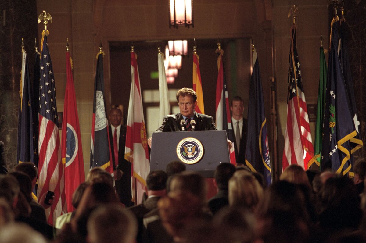 A man stands at a podium on a stage with flags in front of a seated crowd. 
