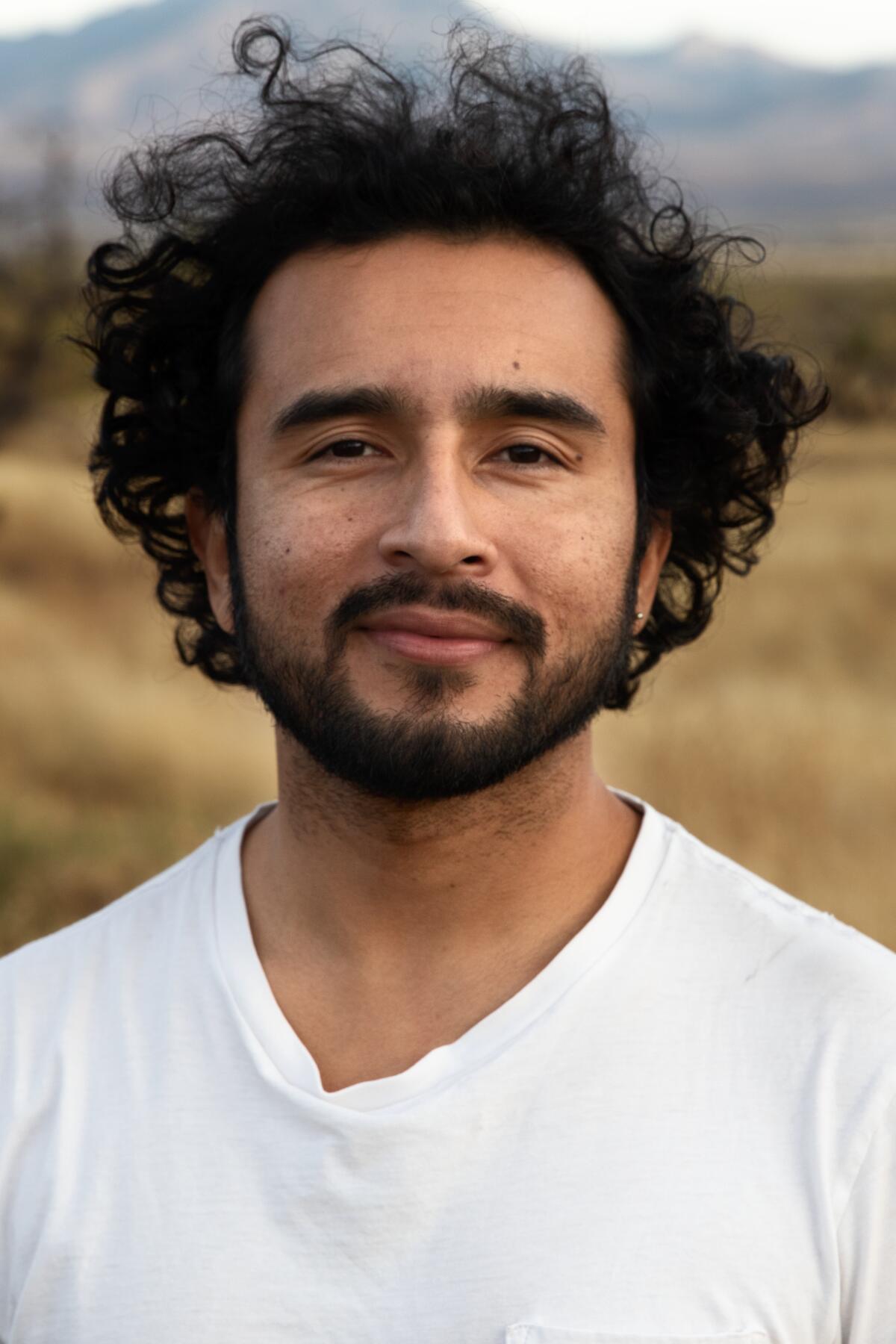 A man with dark, curly hair and a beard, wearing a white T-shirt.