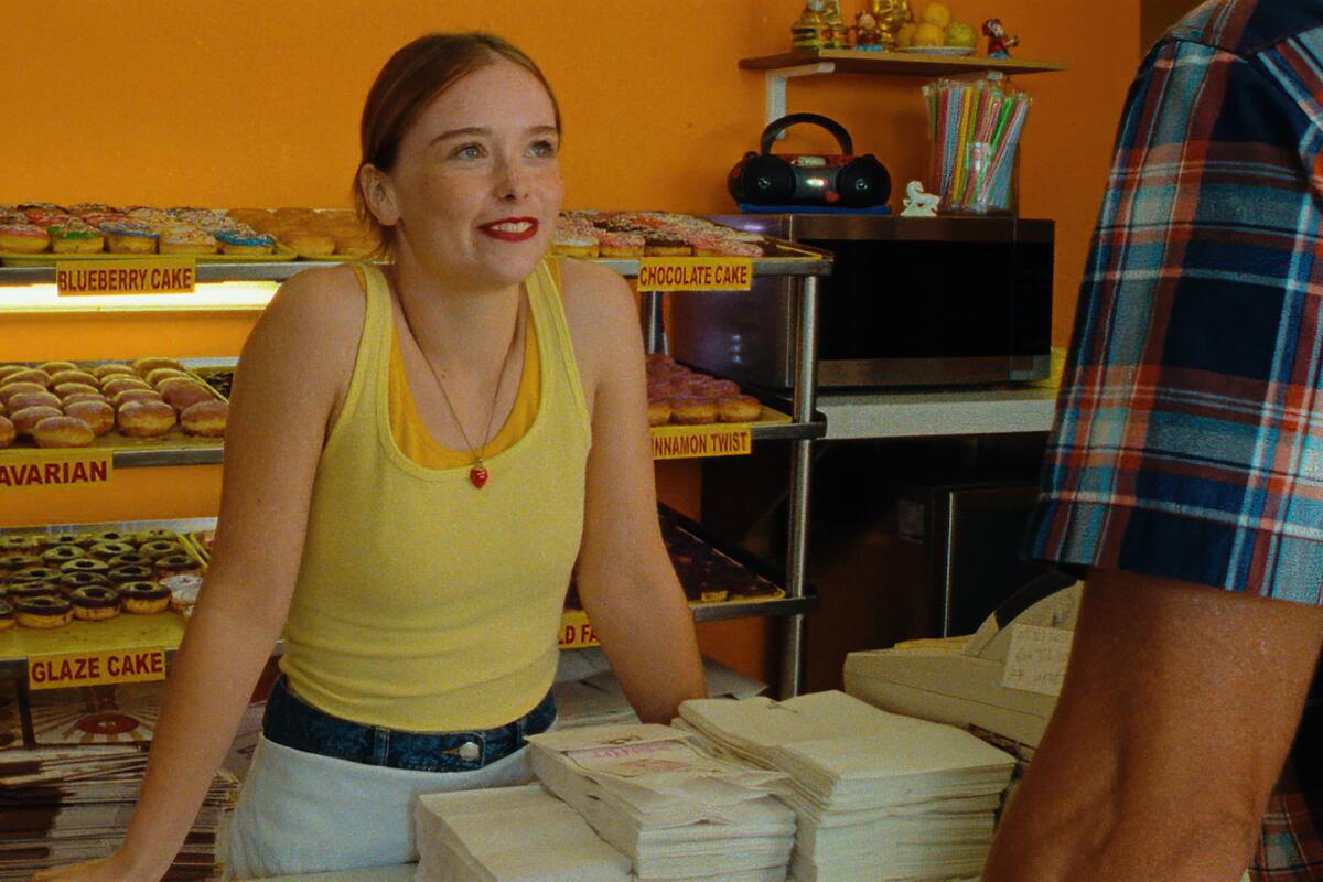 A woman in jeans and a yellow tank top stands behind a counter with racks of doughnuts behind her.