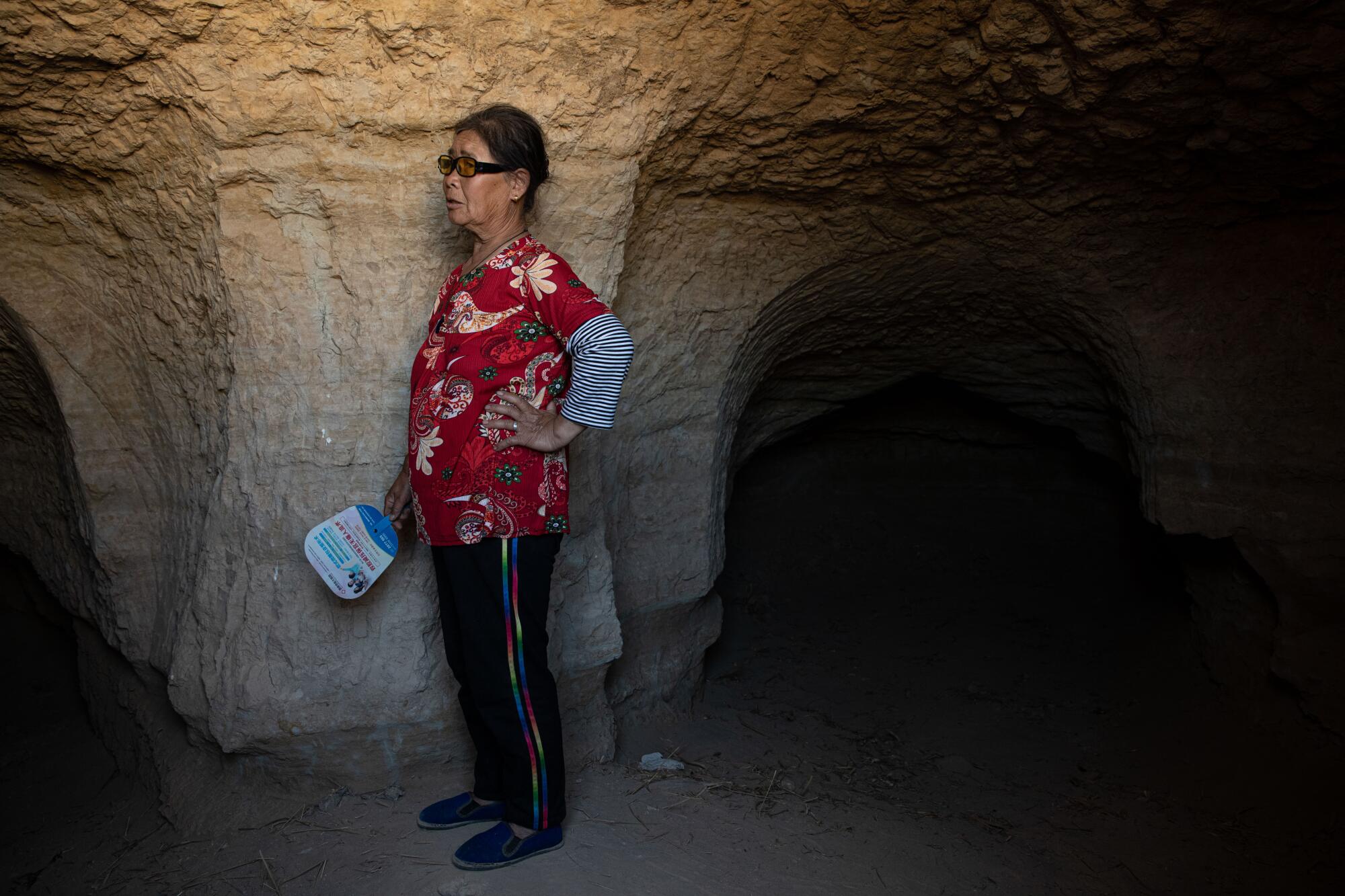 A woman in a red flowery top and dark pants stands near a stone arch in a cave