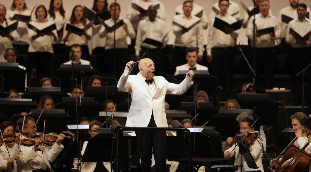 An older man with gray hair and wearing a white suit jacket and black bow tie conducts an orchestra in white.