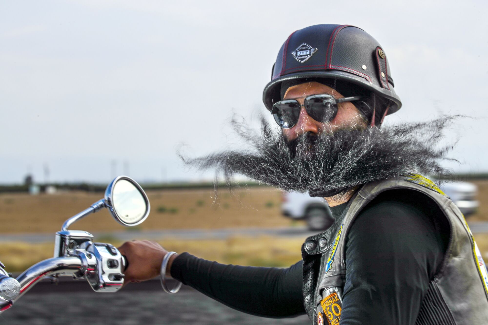 A man, wearing a leather vest and helmet, with a long salt-and-pepper beard looks at the camera while riding a motorbike