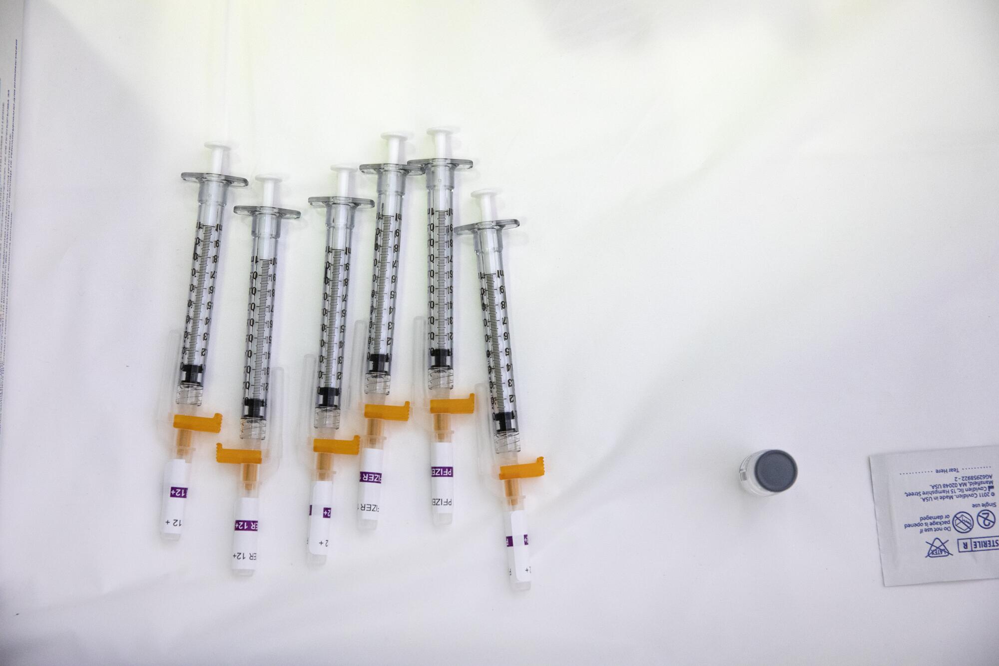 Six syringes of COVID-19 vaccine on a white surface.