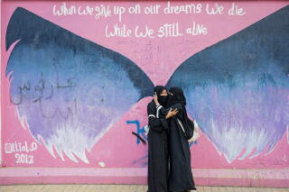 Two women in dark robes and head coverings embrace against a backdrop of blue wings painted on a pink background