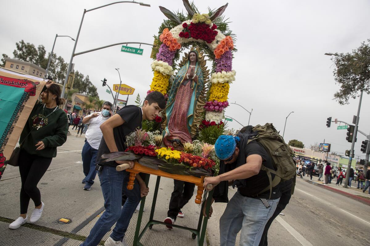 A group of people walks on a street and holds up a large wooden shrine featuring a statue of the Virgen de Guadalupe.