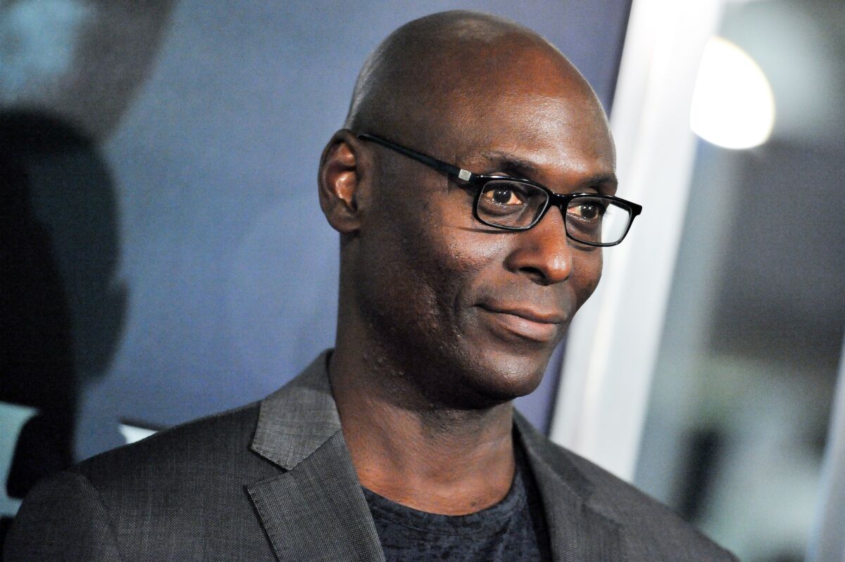 An angled photo of Lance Reddick wearing thin black glasses, a gray blazer and a black shirt