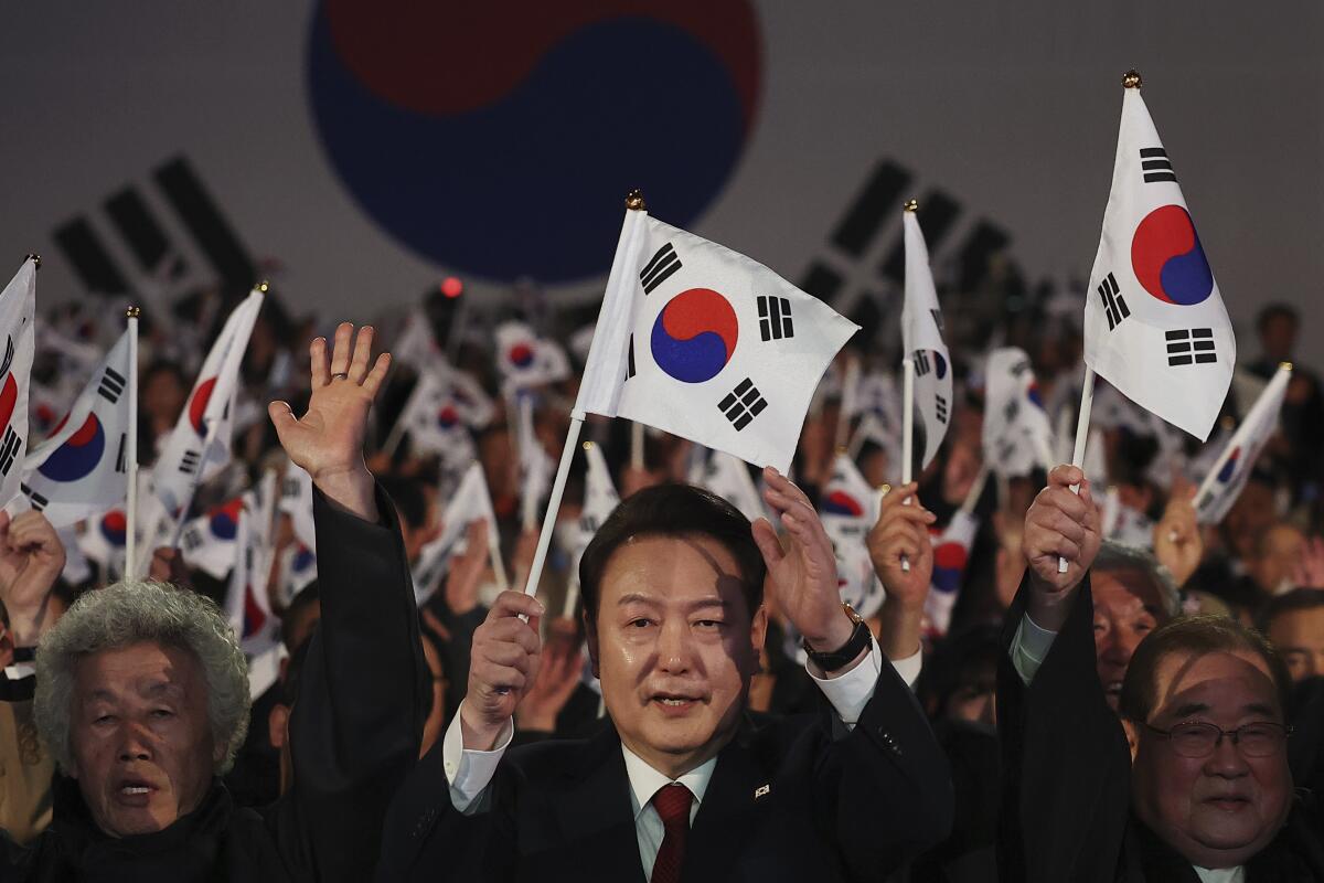 A man in dark suit and red tie holds a white, red and blue flag, surrounded by a crowd of people holding similar flags 