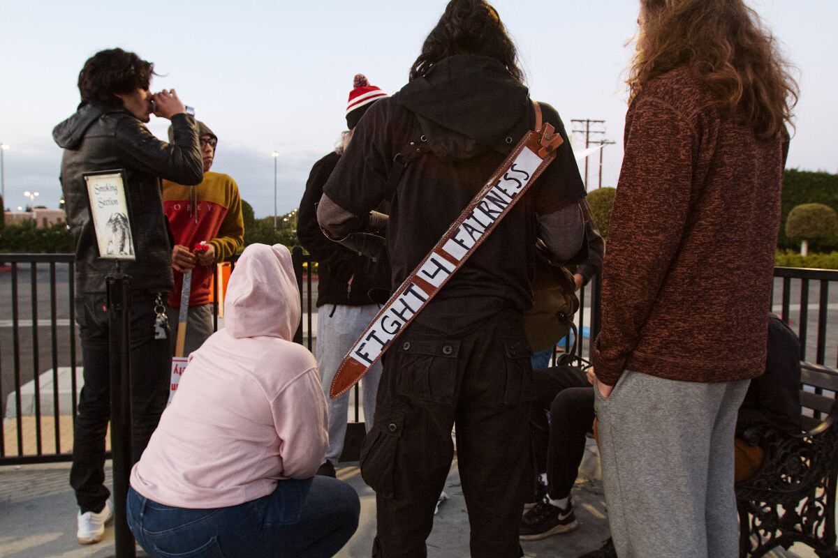 Cast members gather, backs to camera, in the parking lot. A sword sheath reads "FIGHT 4 FAIRNESS" across a person's back.