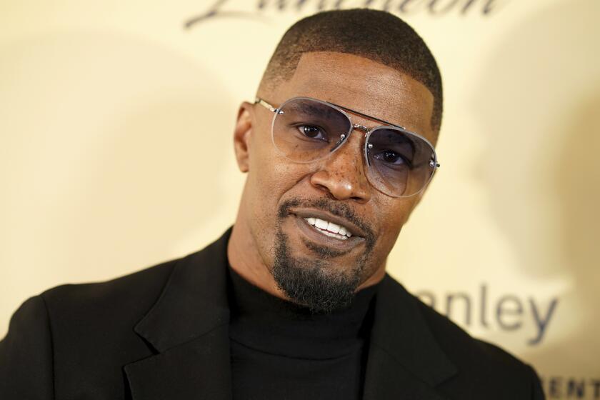 Jamie Foxx smiles and tilts his head while wearing aviator sunglasses and a black shirt and suit