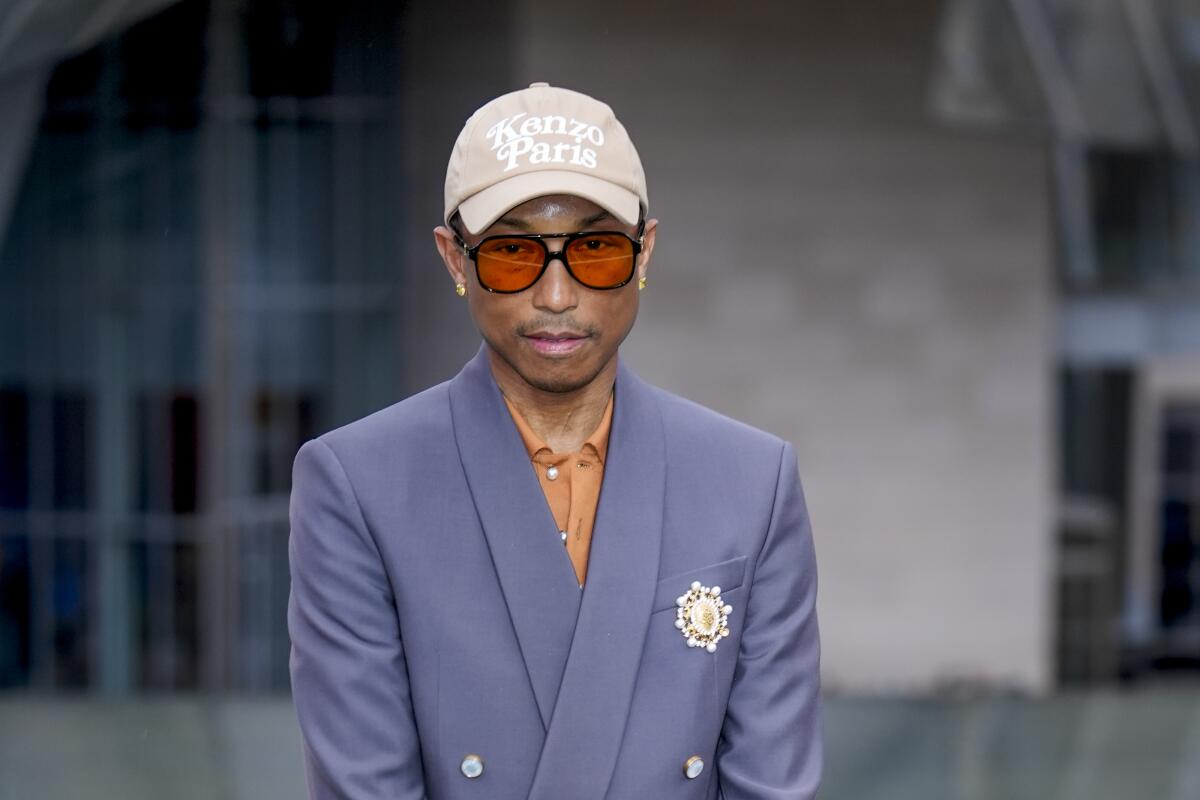 Pharrell Williams posing in a periwinkle suit, orange shirt and tan baseball cap in Paris