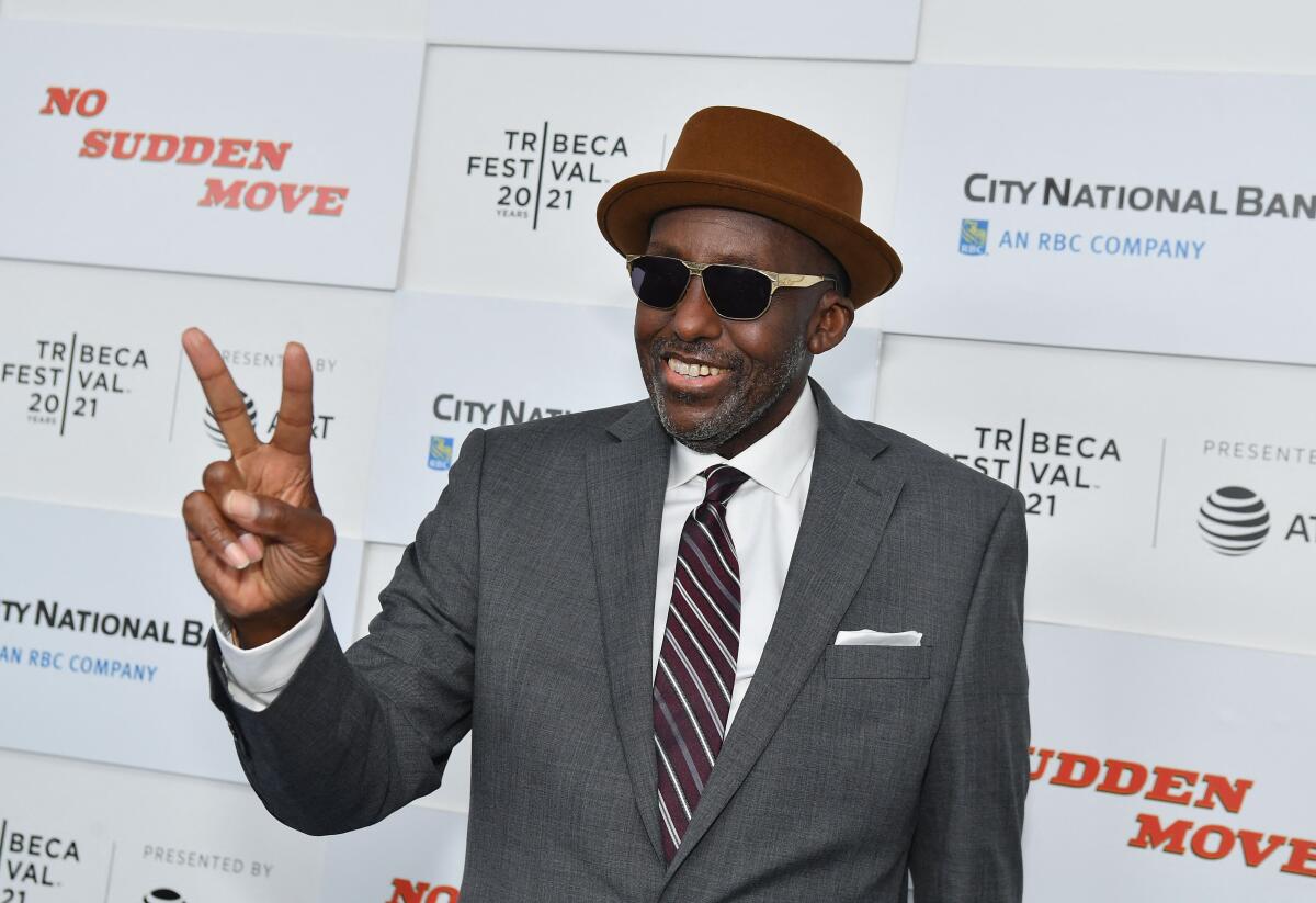 A man wearing sunglasses and a hat makes a peace sign while walking the red carpet at a film festival. 