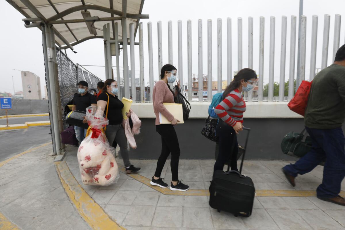 People wearing masks and carrying belongings walk by a fence