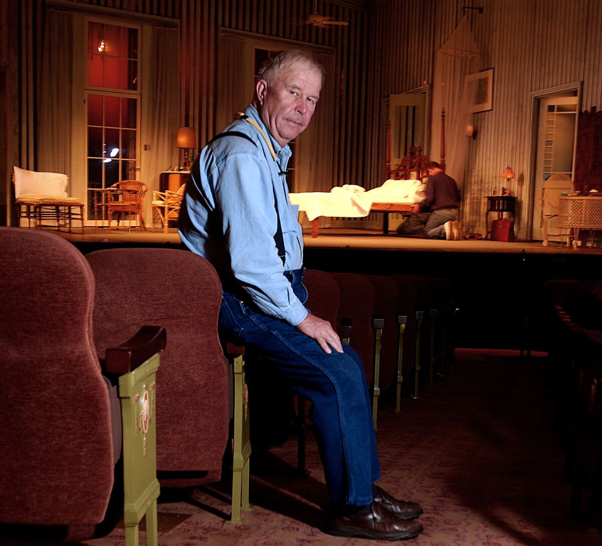 Ned Beatty perches on the arm of a theater seat in front of a stage dressed for a play.