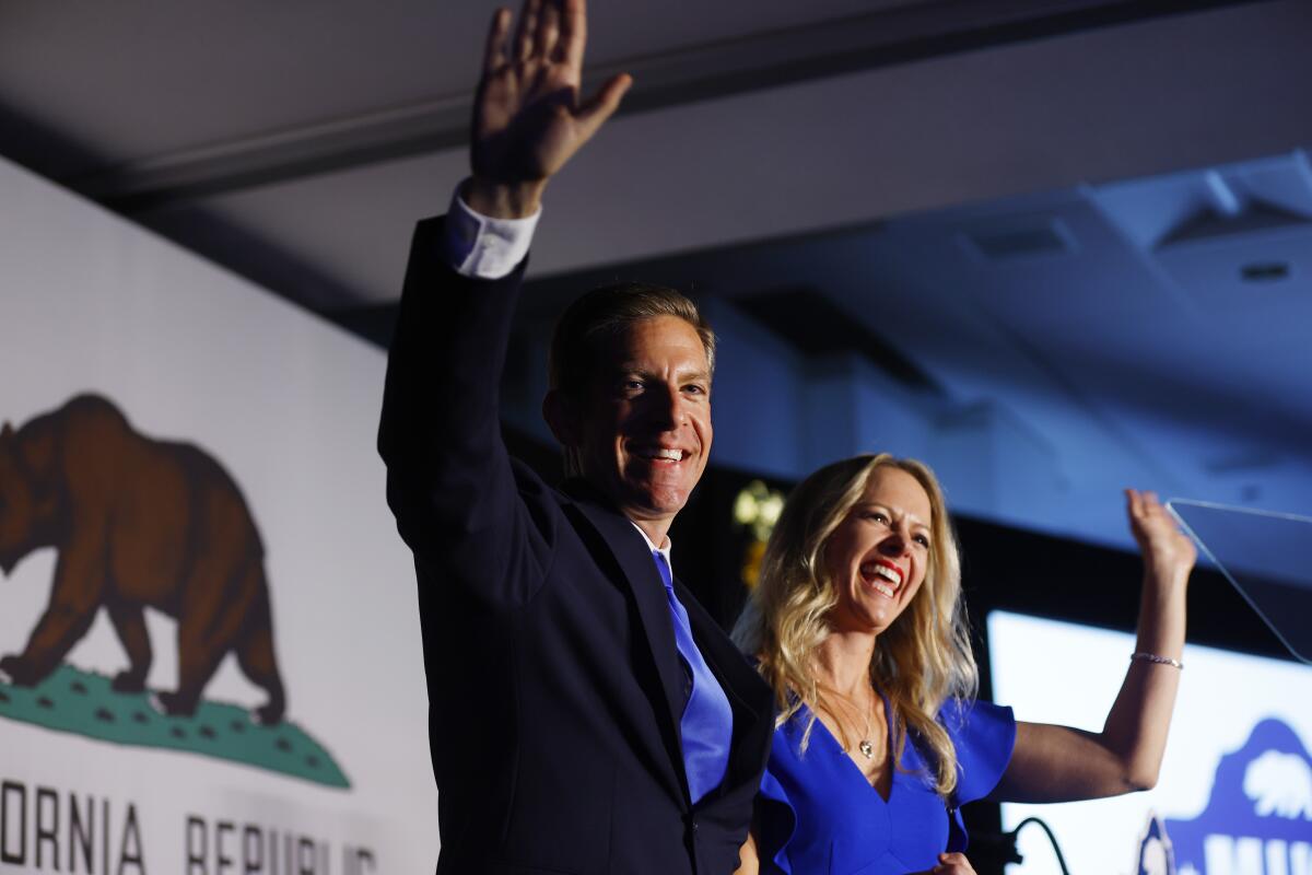 A man and woman, smiling, wave from a stage