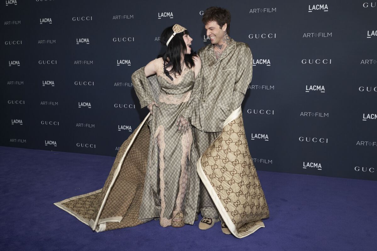 A man and a woman in matching brown outfits share a big blanket and smile at each other.