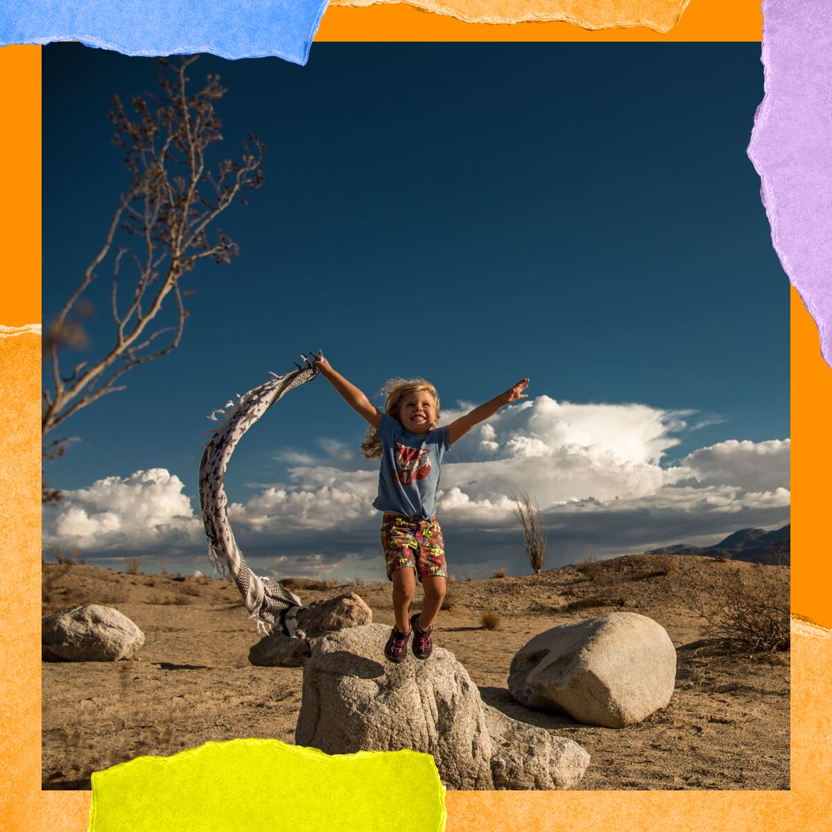 A child jumps of a desert rock gleefully while holding a piece of fabric that blows in the wind.