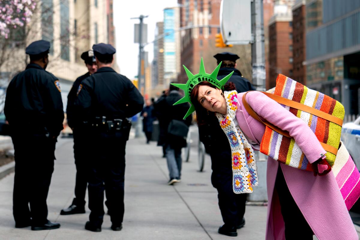 A woman wearing a pink coat and a Statue of Liberty hat bent over slightly on a New York sidewalk