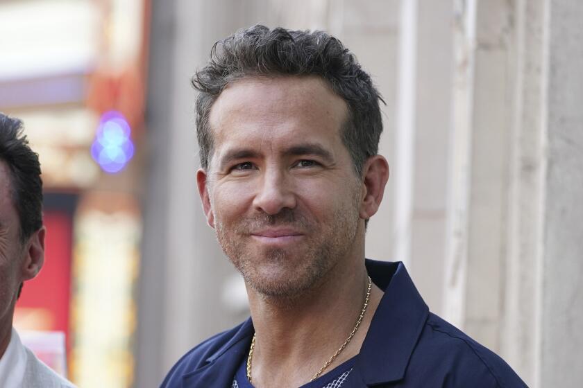 Ryan Reynolds grinning with no teeth, wearing a dark blue shirt. 