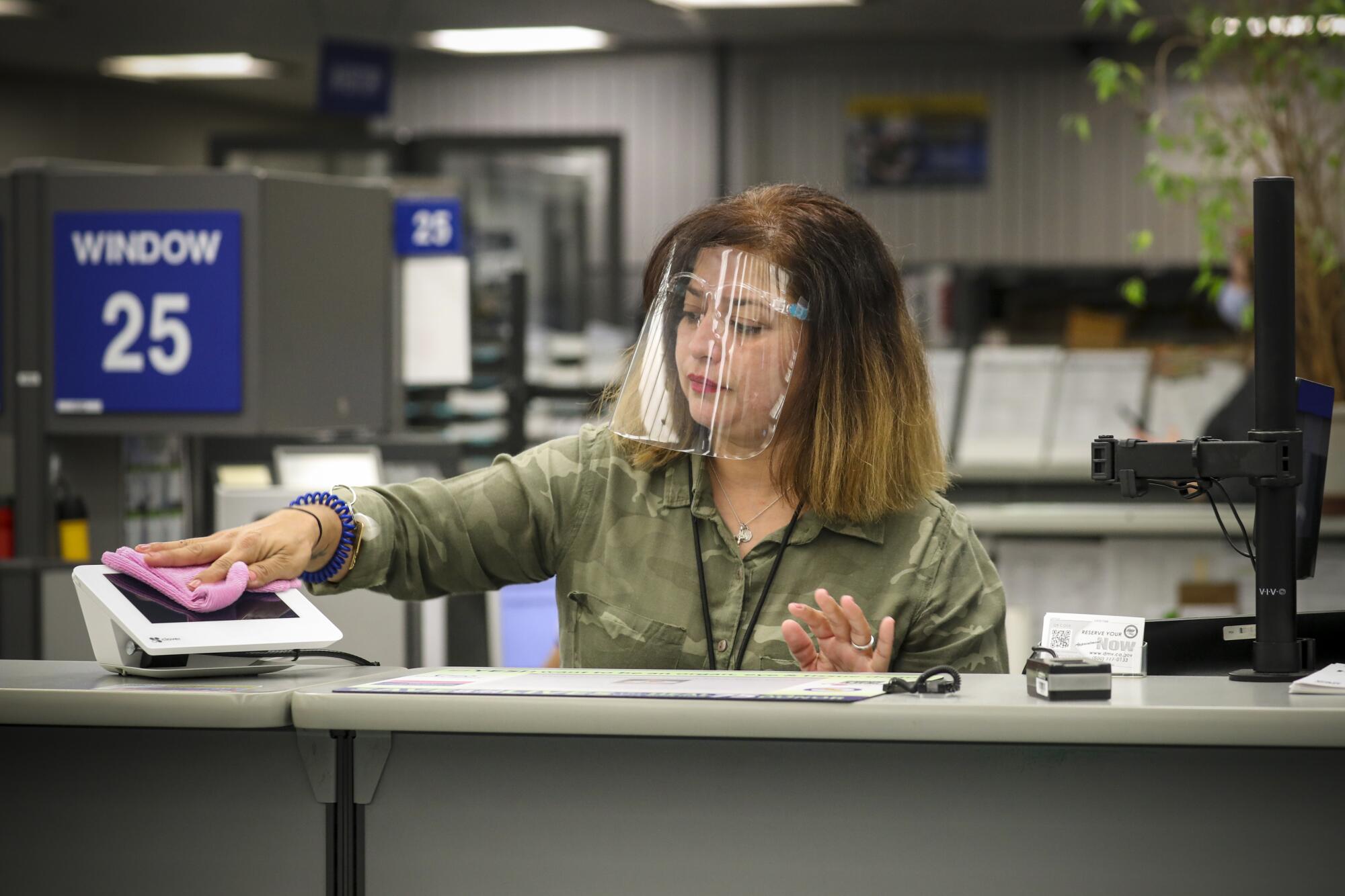 A DMV worker wearing a transparent plastic face shield wipes down an electronic terminal at her workstation